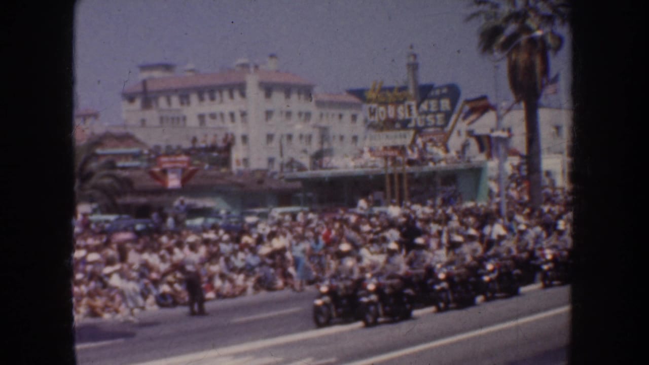 Motorcycle parade passes a crowded sidewalk near palm trees and a large venue sign.