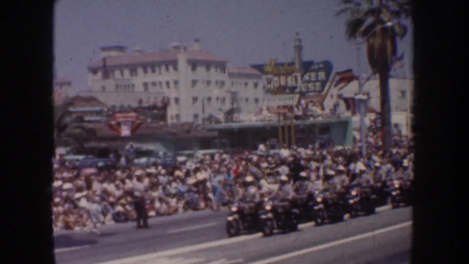 Motorcycle riders move along a California city street in 1952 as spectators crowd the sidewalk near palm trees, commercial buildings, and a large venue sign.