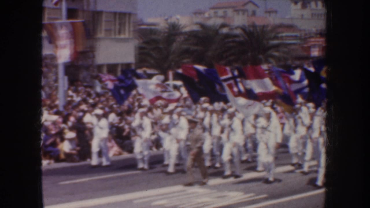 1952 California home-movie frame showing sailors in white uniforms marching with colorful flags before a crowd and palm-lined buildings.