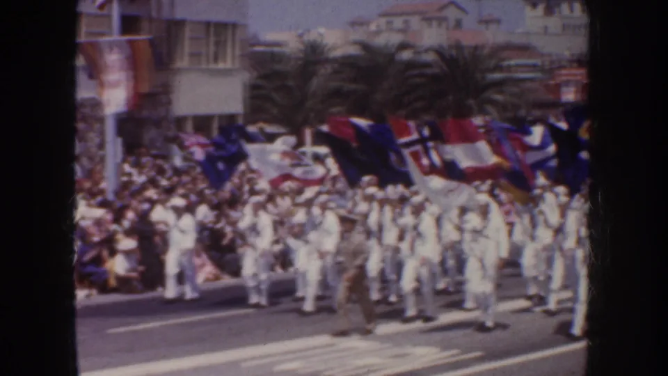 1952 California home-movie frame showing sailors in white uniforms marching with colorful flags before a crowd and palm-lined buildings.