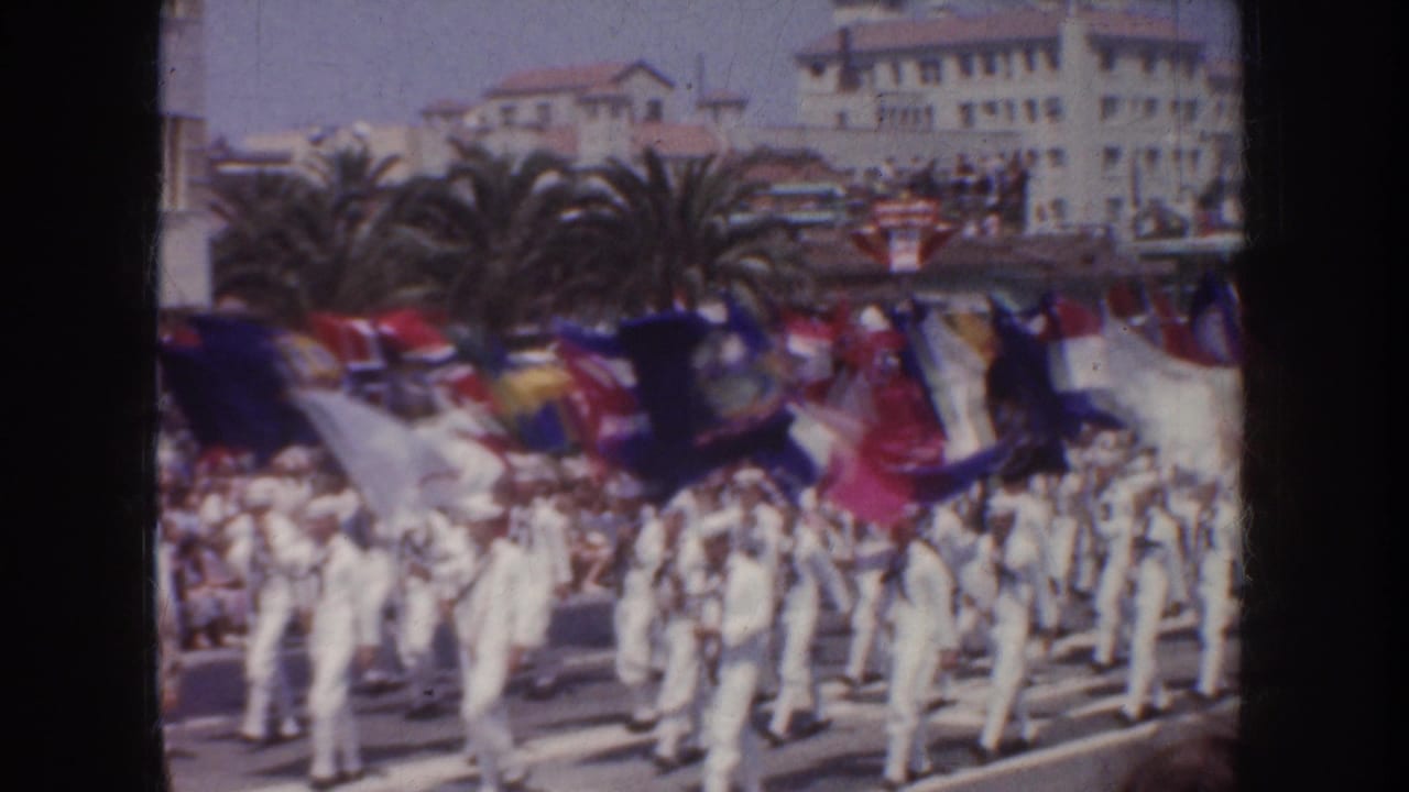 A formation of white-uniformed sailors carries flags along a street with crowds behind them.