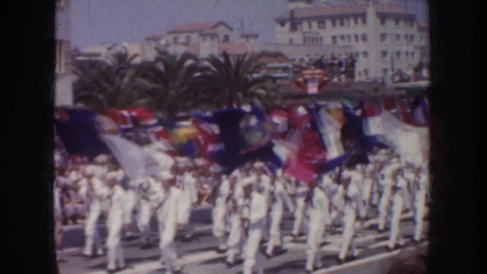 1952 California parade view with sailors marching in formation, vivid flags overhead, spectators and palm trees in the background.