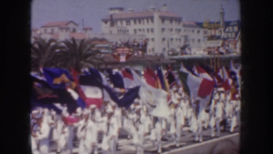 1952 California home-movie frame showing a dense cluster of parade flags, white-uniformed marchers, crowds and mid-century street architecture.