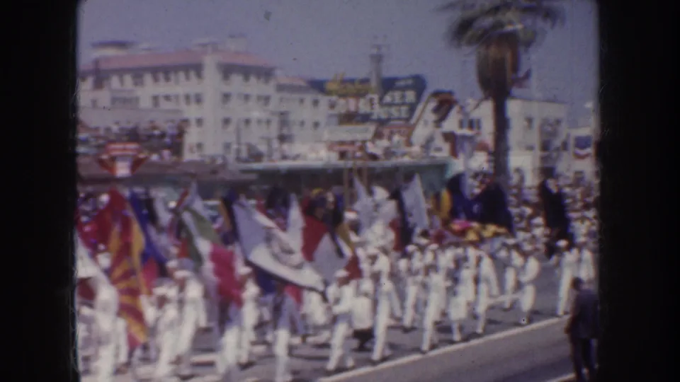 1952 California parade frame with sailors, vivid flags, spectators, a palm tree and a possible fountain or sign structure along the route.