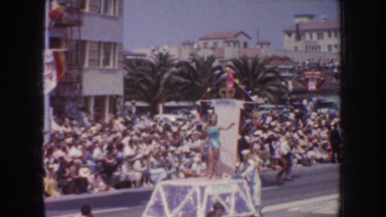 1952 California parade frame showing a woman in a light swimsuit on a decorated float, waving toward spectators along a palm-lined street.