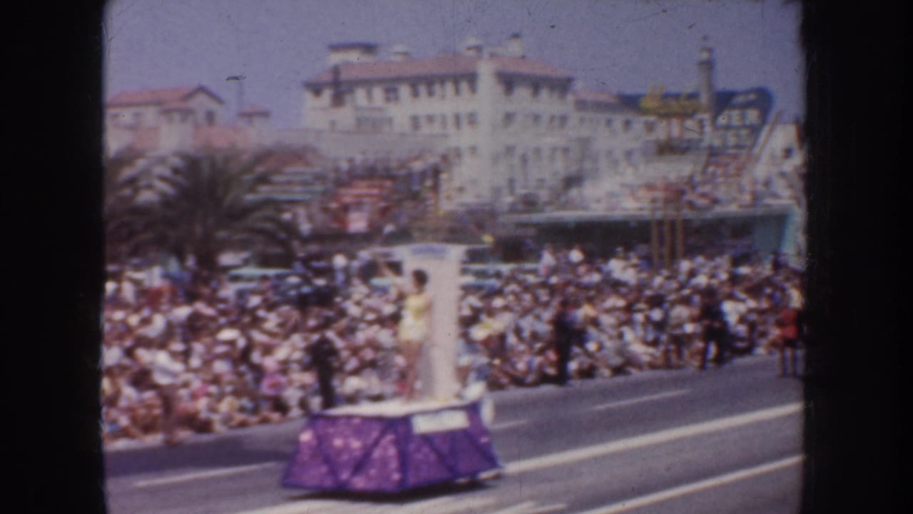 A purple parade float passes a dense crowd with buildings and palm trees behind it.
