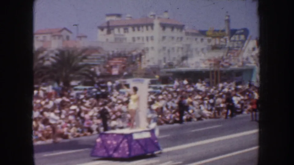 1952 California parade view of a purple float moving past spectators, palm trees, and mid-century streetfront buildings.