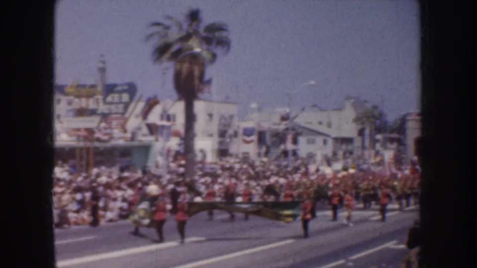 1952 California street parade frame with marching band members carrying a banner or formation piece before a packed sidewalk crowd.