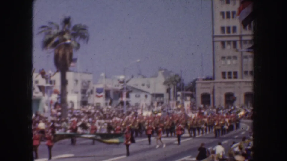 1952 California parade frame showing a marching band moving past a tall palm, bunting, storefronts, and a dense crowd of spectators.