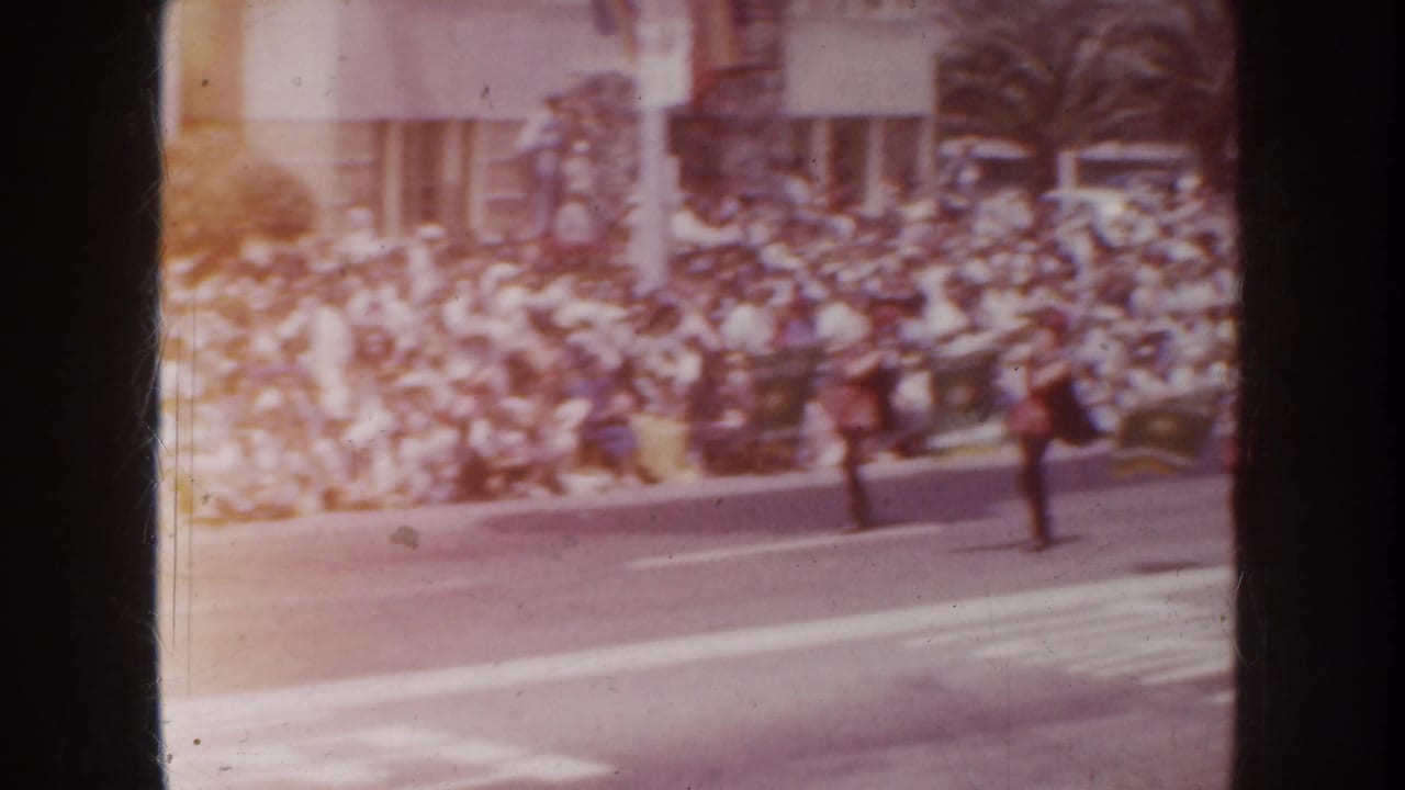1952 California home-movie frame showing a large curbside parade crowd with uniformed figures crossing the street.