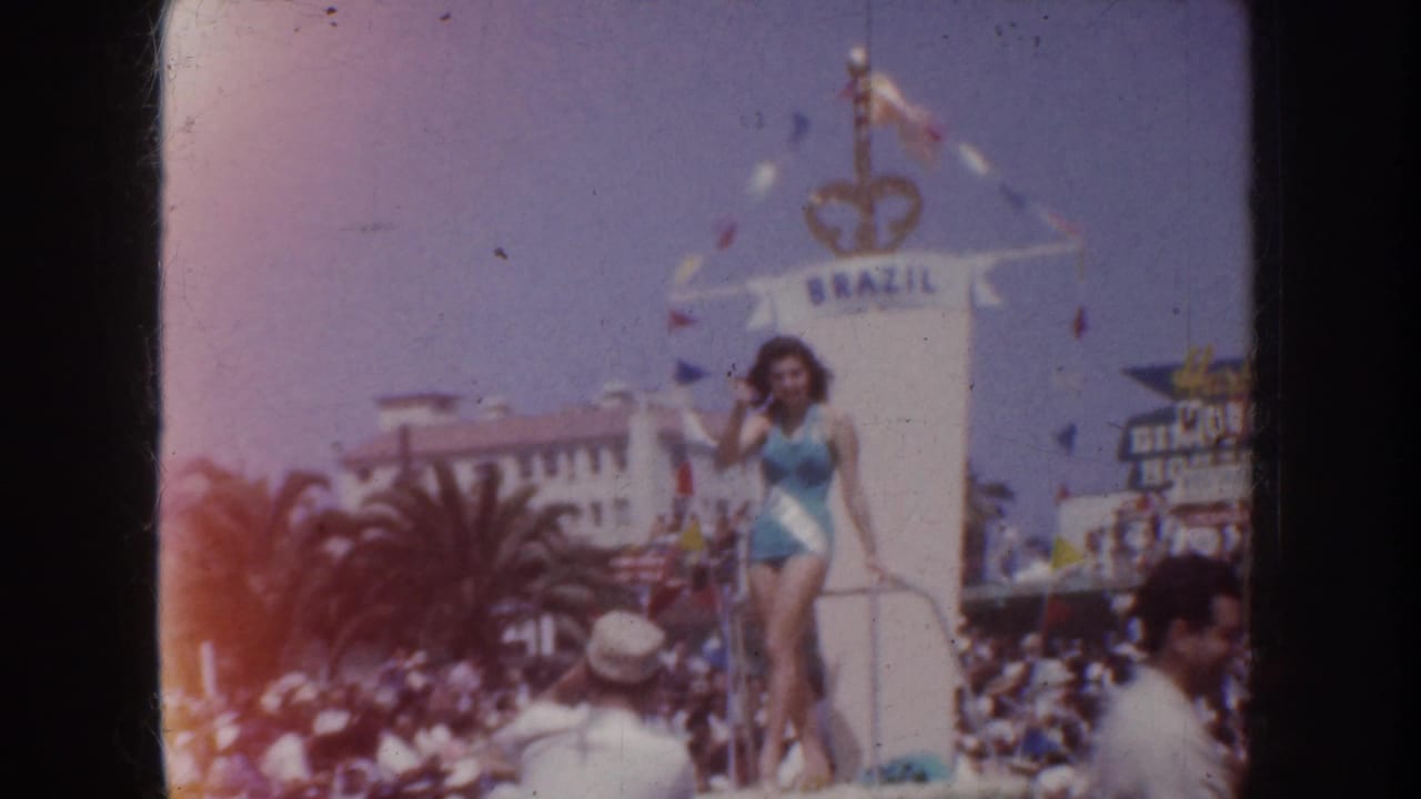 Woman in a turquoise swimsuit waves from a Brazil-labeled parade float before a crowd.