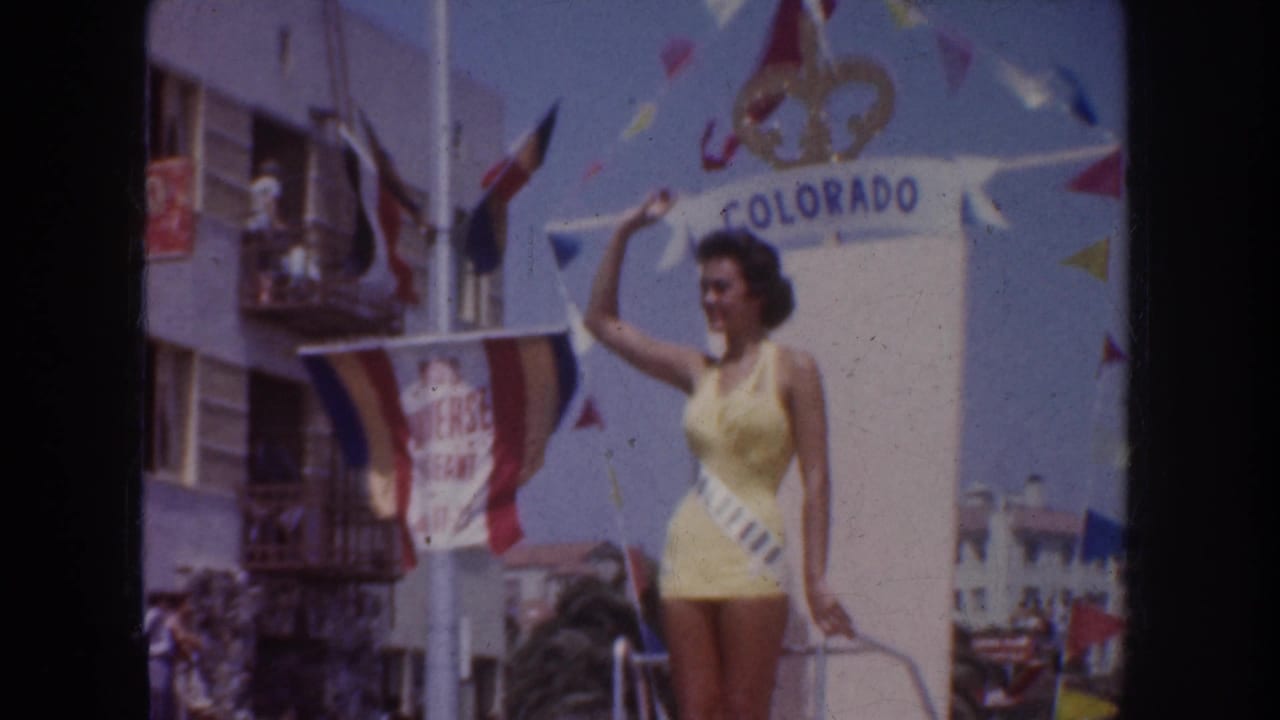 Contestant in yellow stands beside a Colorado sign on a parade float near buildings and flags.