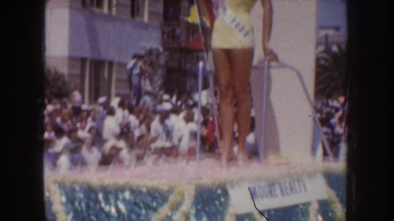 Home-movie close framing shows the lower portion of a pageant contestant on a decorated float, with a crowd behind her.