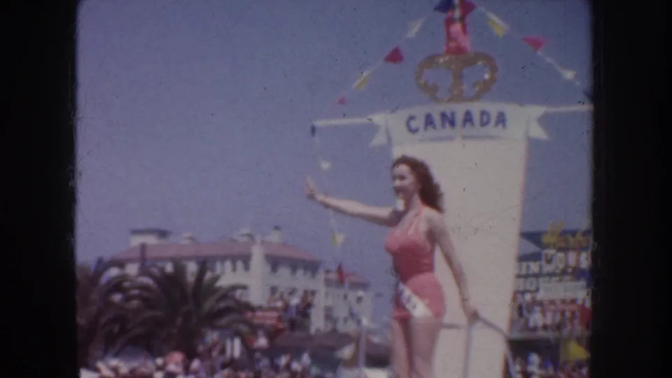 1952 California parade frame showing a contestant on a Canada-labeled float, waving above a crowd and palm-lined street.