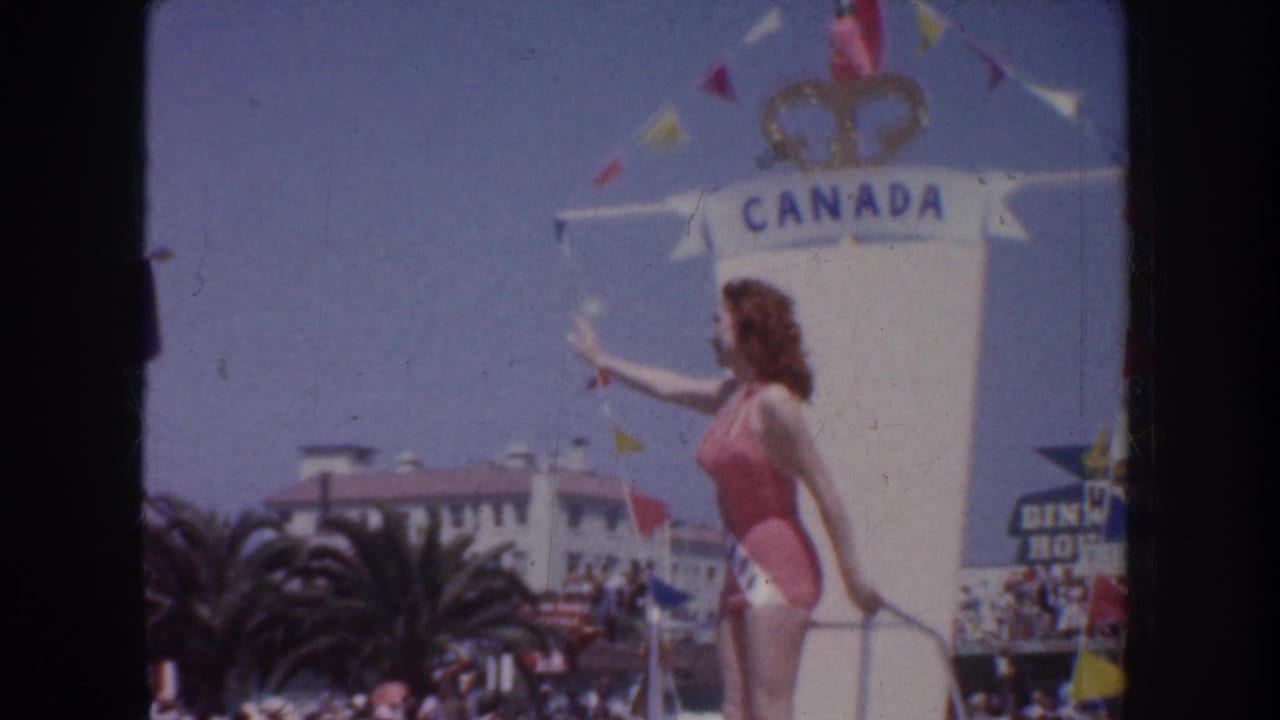 1952 California home-movie frame showing a sash-wearing beauty queen waving from a float near a large Canada sign and pennant flags.