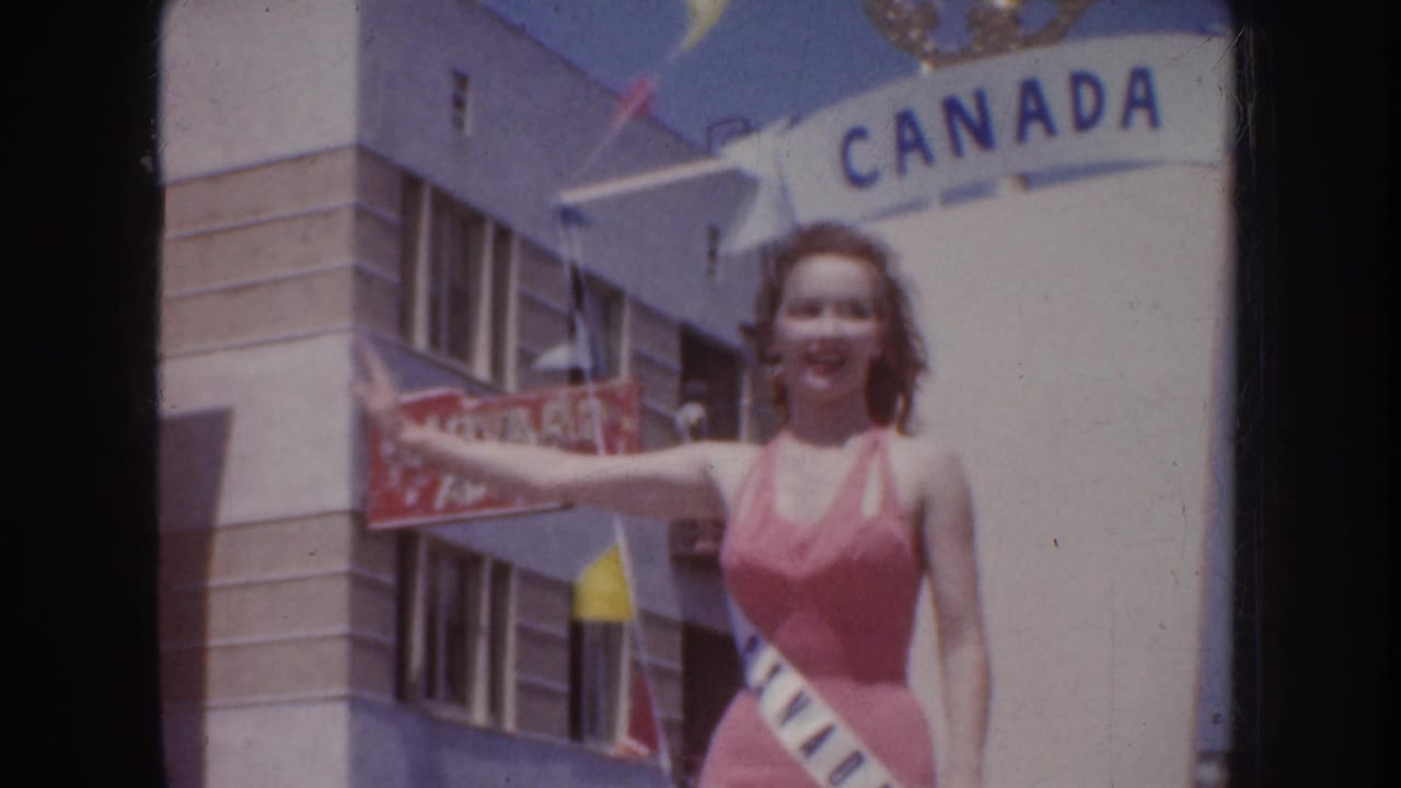 Smiling beauty queen in a sash stands near a Canada sign with a building and flags behind her.