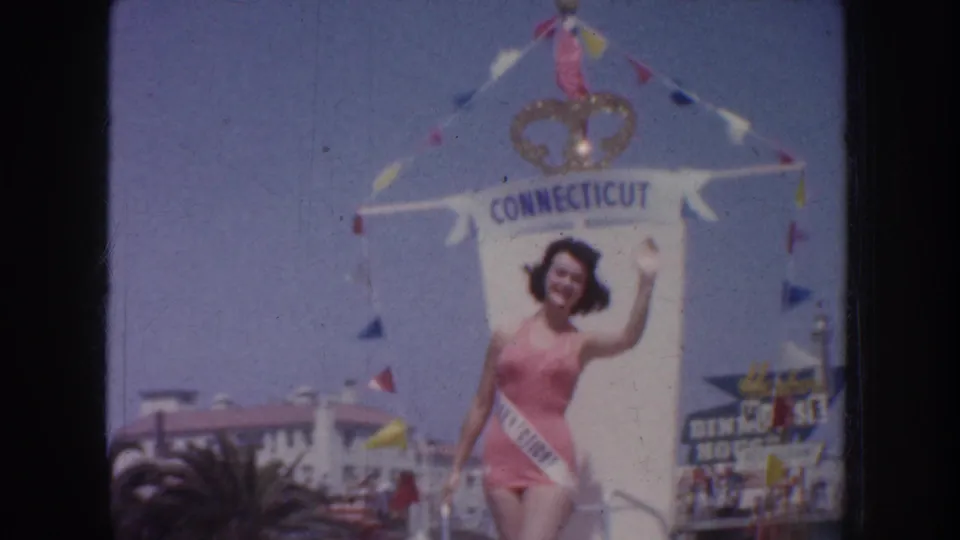 1952 California home-movie frame showing a waving contestant on a float labeled Connecticut, with flags and street spectators visible.