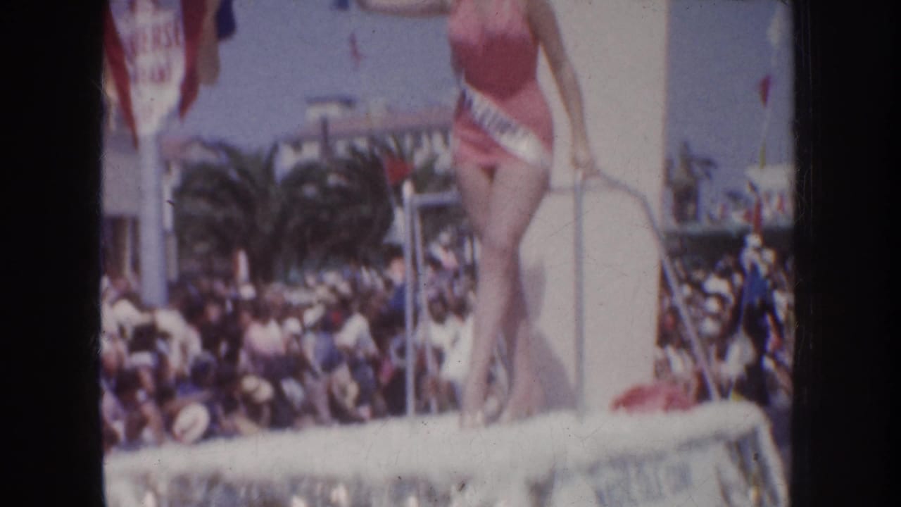 1952 California parade frame showing a sash-wearing beauty queen on a decorated float, with spectators, palm trees, flags, and signage behind her.