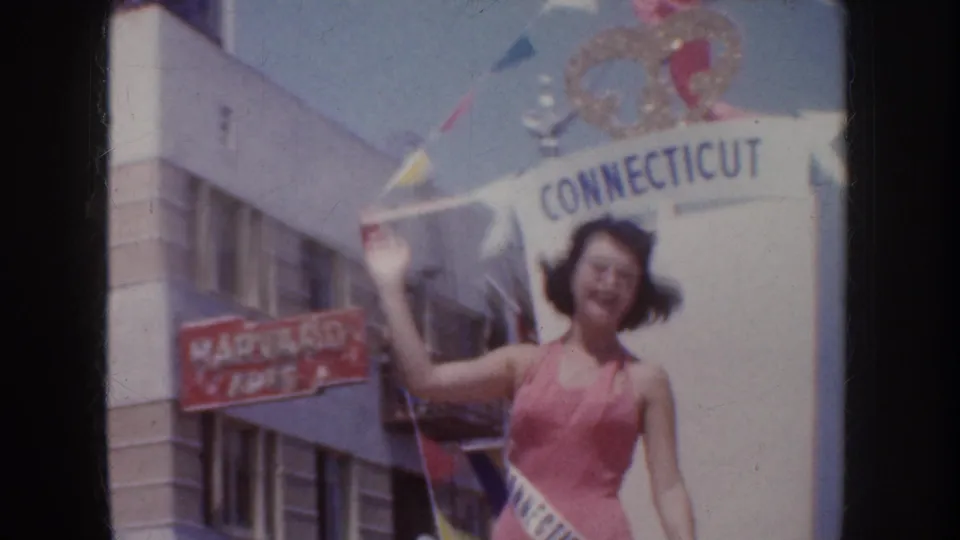 1952 California parade frame showing a sash-wearing woman in pink waving near a Connecticut banner and storefront signage.