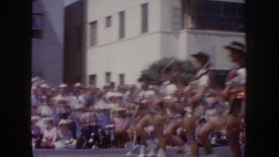 Parade dancers in matching outfits move past seated spectators and mid-century buildings in 1952 California home-movie footage.