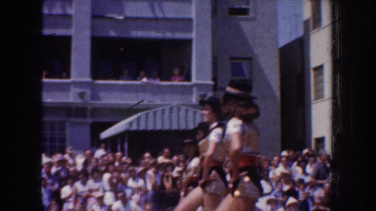 A handheld 1952 California parade view shows dancers passing a crowded sidewalk and people watching from upper balconies.