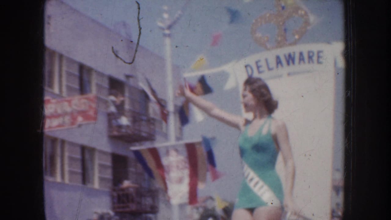 Woman in a teal swimsuit and Delaware sash waves beside parade float lettering.