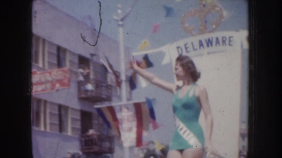A woman in a teal swimsuit with a Delaware sash waves from a parade float in 1952 California footage.