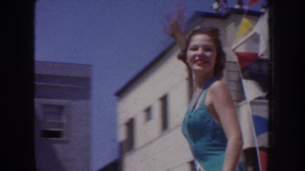 A smiling swimsuit contestant waves from the float area, framed by building walls and colored parade decorations.