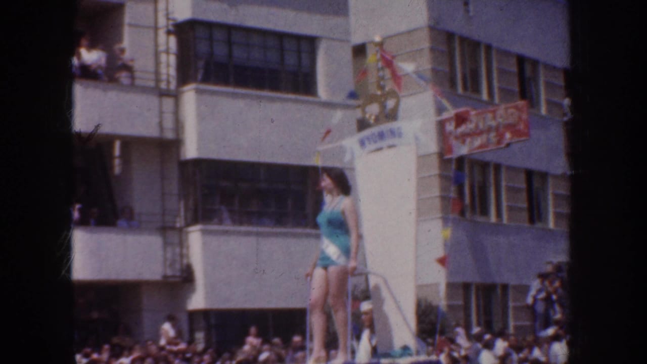 1952 California parade frame showing a woman in a turquoise swimsuit on a float, with spectators, banners, and commercial buildings behind her.