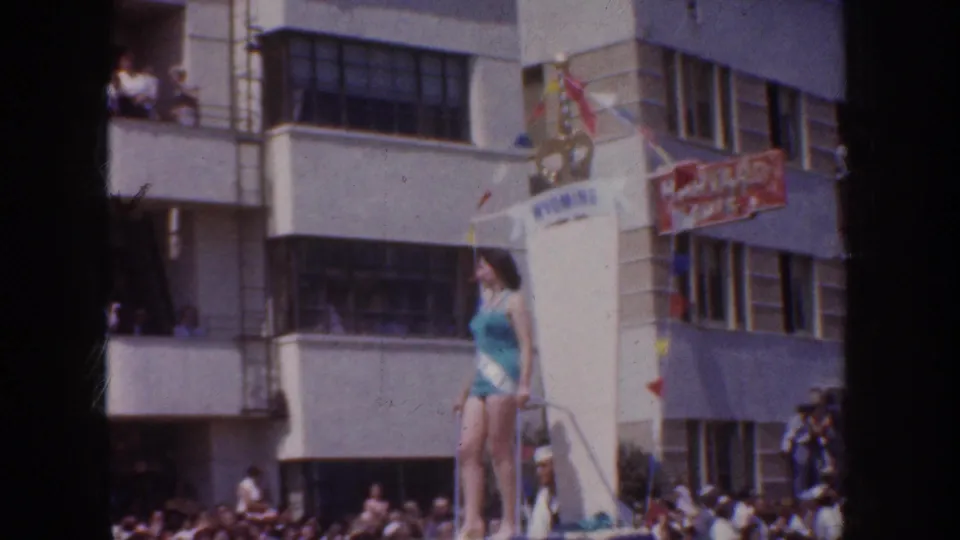 1952 California parade frame showing a woman in a turquoise swimsuit on a float, with spectators, banners, and commercial buildings behind her.