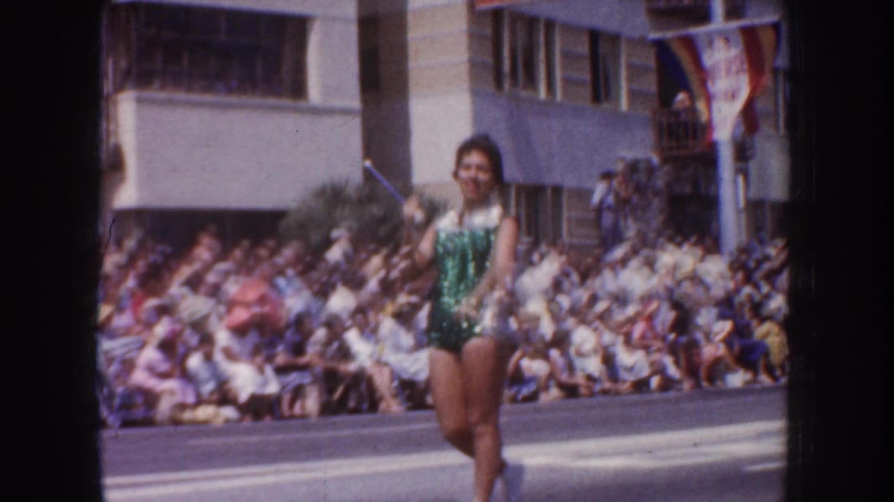 1952 California parade frame showing a woman in a green sequined outfit performing near the curbside crowd and buildings.