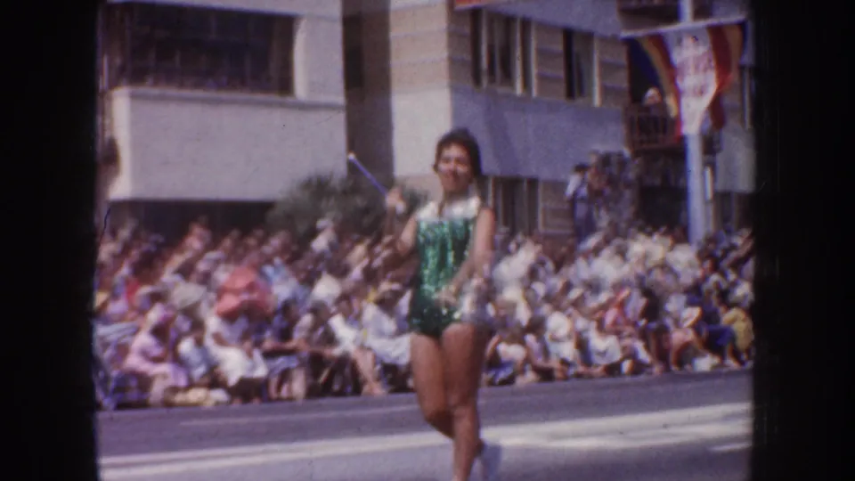 1952 California parade frame showing a woman in a green sequined outfit performing near the curbside crowd and buildings.
