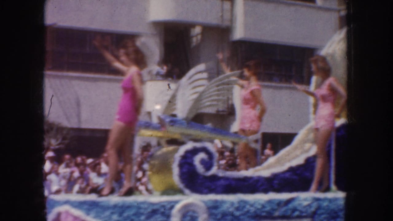 Two women in swimsuits wave from a decorated parade float with a large seahorse form.