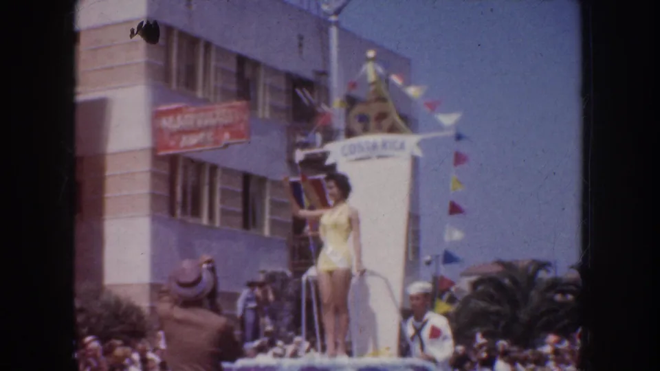 1952 California 8mm frame showing a woman in a yellow swimsuit on a COSTA RICA parade float with pennants, spectators, and a HARVARD sign nearby.