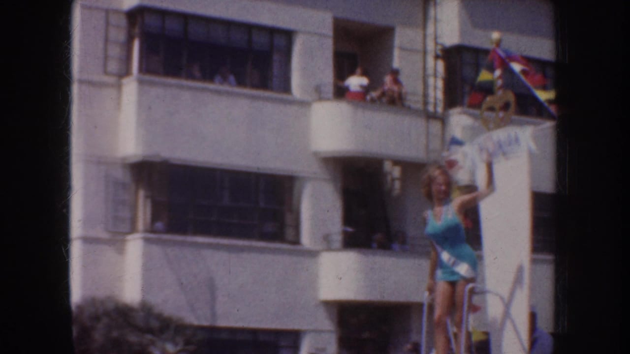 Woman in a blue swimsuit stands on a parade float below balcony spectators.