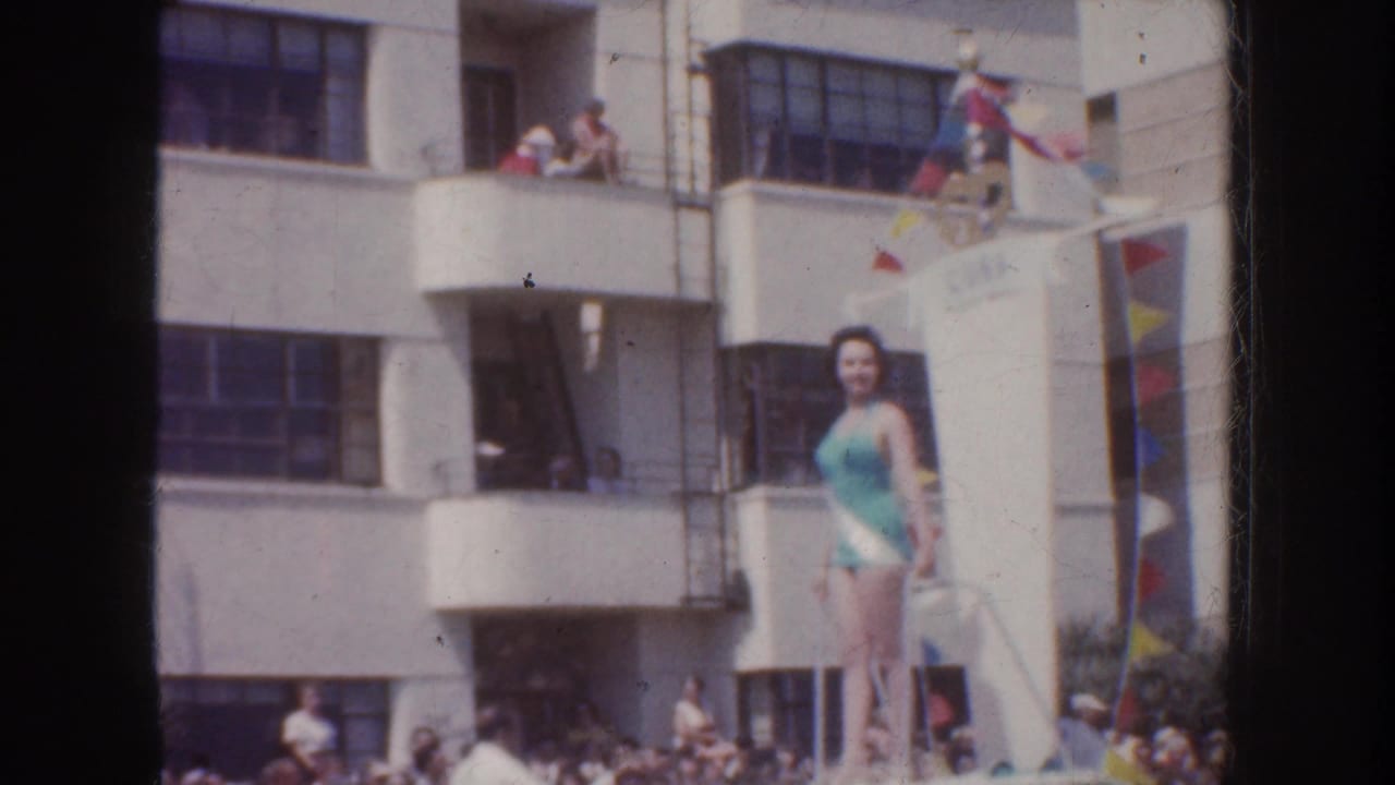 1952 California 8mm frame showing a blue-swimsuit parade rider on a float, with spectators in balconies and along the street.