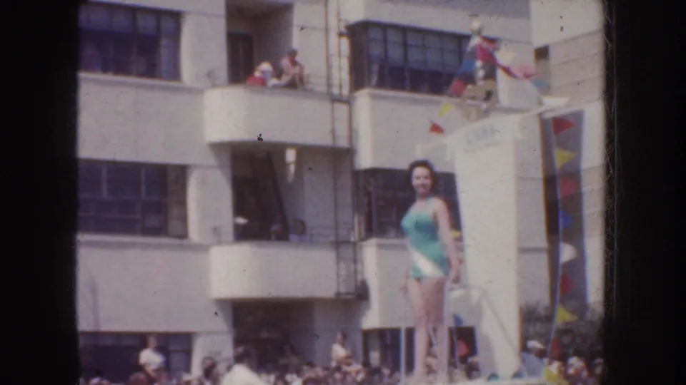 1952 California 8mm frame showing a blue-swimsuit parade rider on a float, with spectators in balconies and along the street.