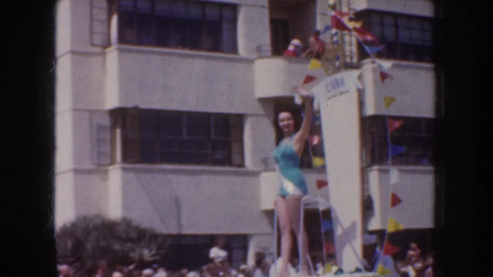1952 California parade frame with a woman in a blue swimsuit waving from a decorated float below balcony spectators and colorful pennants.