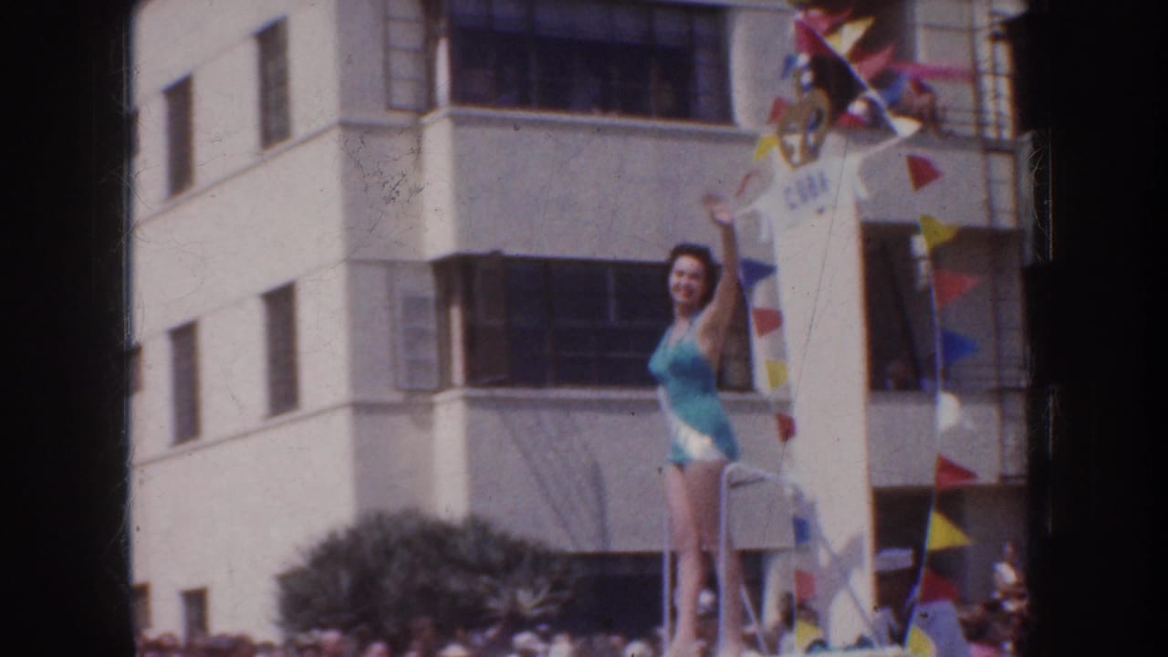 In 1952 California parade footage, a woman in a turquoise one-piece swimsuit waves from an elevated decorated float as spectators gather below.
