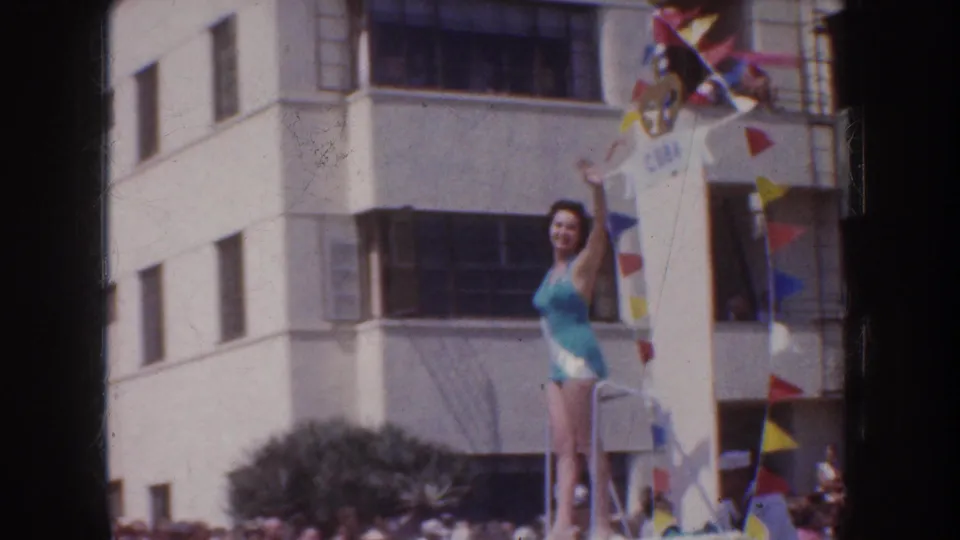 In 1952 California parade footage, a woman in a turquoise one-piece swimsuit waves from an elevated decorated float as spectators gather below.