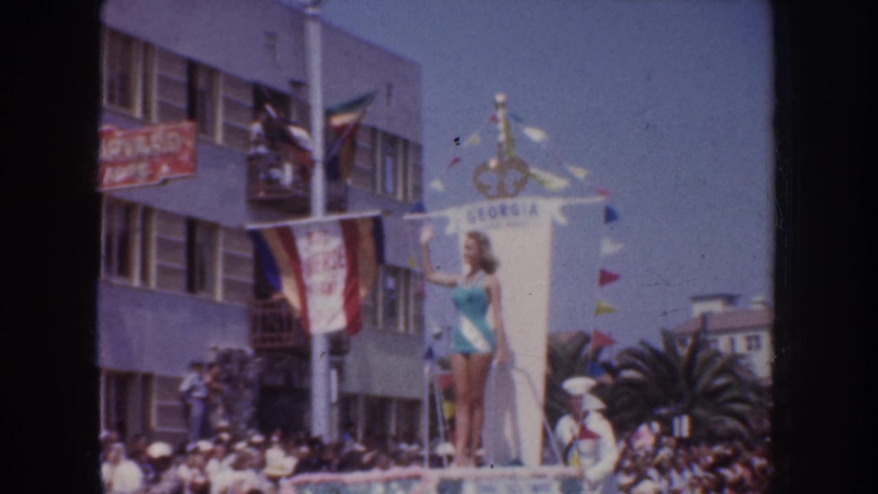 Woman stands on a GEORGIA-labeled parade float with flags, crowd, palm tree, and building balconies behind her.