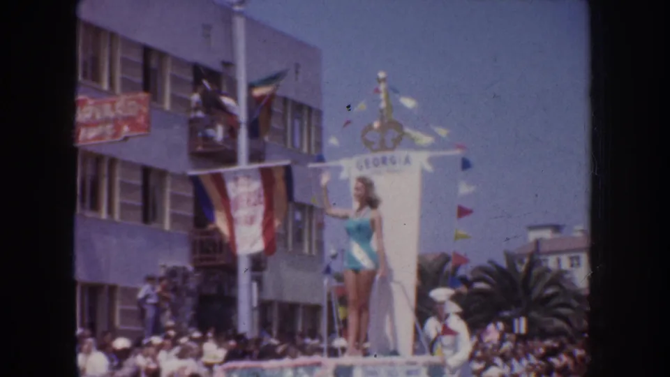 A woman stands on a GEORGIA-labeled parade float during a 1952 California civic parade, with flags, palm trees, and spectators behind her.