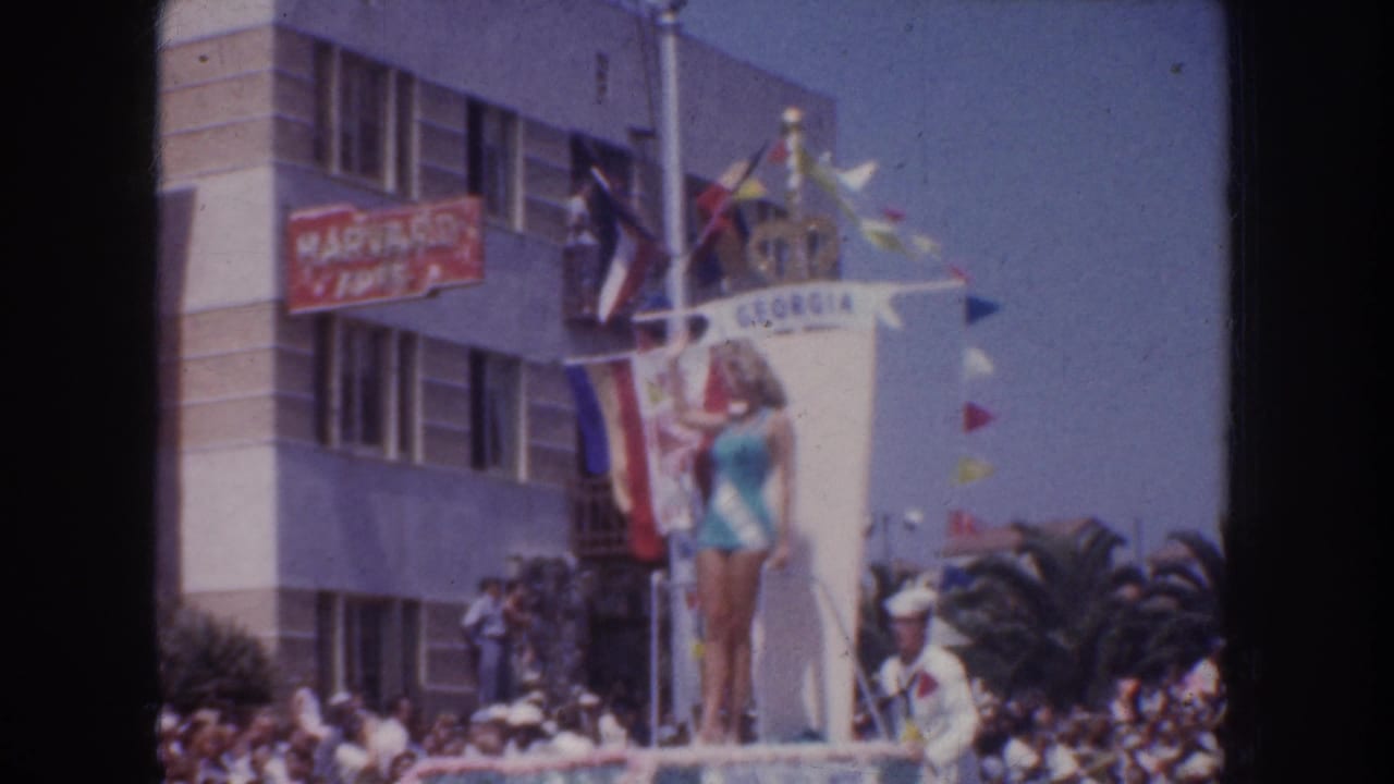 The GEORGIA parade float is seen from the side, with the woman posed above the crowd and uniformed parade staff visible below.