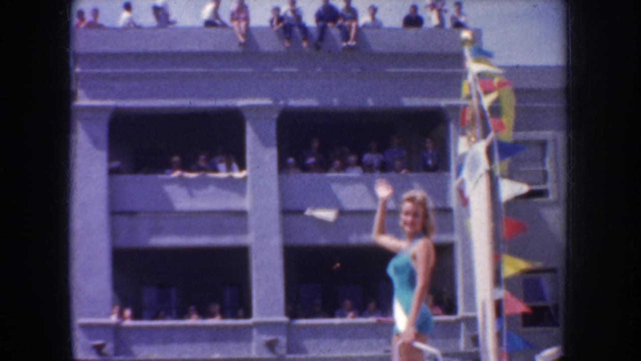 A woman waves from a decorated parade float while spectators watch from balconies, open windows, and the roofline of a nearby building.