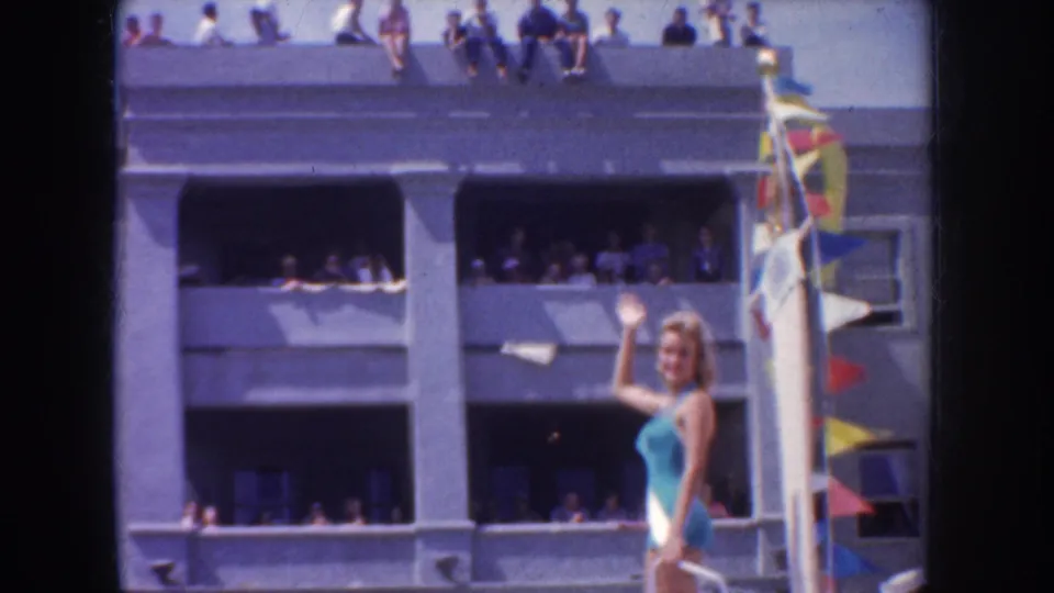 A woman waves from a decorated parade float while spectators watch from balconies, open windows, and the roofline of a nearby building.