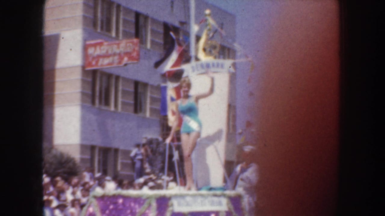 1952 California parade view of a pageant winner on a DENMARK float, with spectators, flags, and city buildings behind her.
