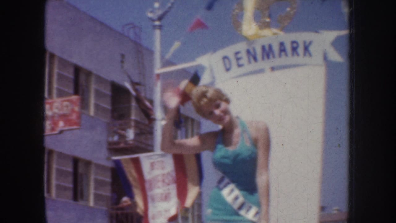 Close view of sash-wearing woman waving beside a DENMARK sign on a parade float.