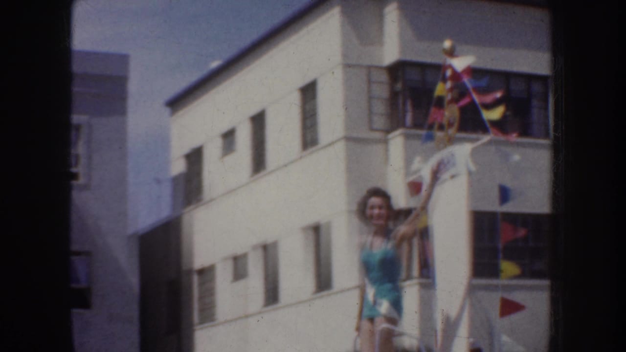 Wide 1952 California parade frame showing the pageant participant on the float with flags, white trim, and urban buildings.
