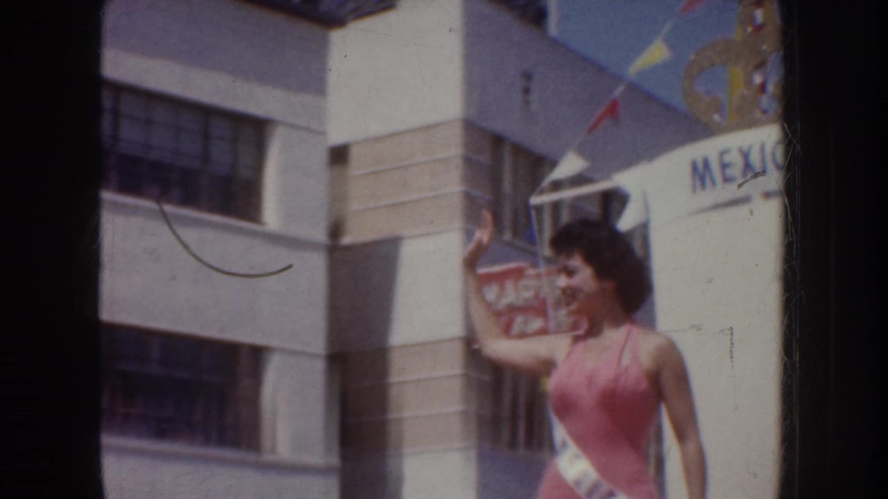 1952 California 8mm frame showing a sash-wearing woman in a pink swimsuit waving near a Mexico sign and pennant flags.