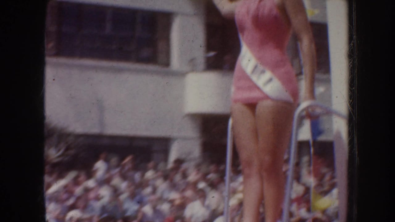 Sash-wearing woman stands near a railing with a blurred crowd behind her.